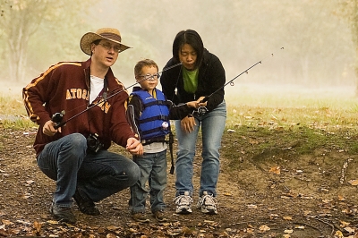 A couple helps a small toddler with their fishing rod on the banks of a pond