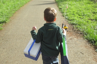 Young boy walking on trail withe arms full carrying cooler, camp chair and fishing rod.