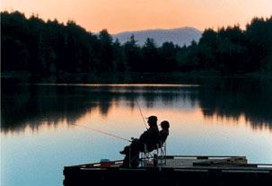 Two people sit in folding chairs on a deck that extends into Coffenbury Lake. The people are holding fishing rods and the sun is setting so that the sky is orange and the people are in silhouette