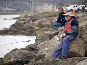 Two people sit on large boulders on the shore of Yaquina Bay with fishing lines in the water