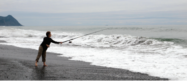 A woman stands barefoot on the beach and casts a long rod into the waves. She is fishing for surfperch.